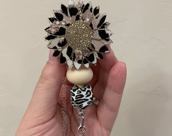 May include: A gold glitter sunflower with a black and white leopard print design. The sunflower is attached to a clear badge holder with a brown and white leopard print bead and a cream colored bead.