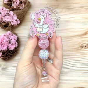 May include: A pink and white badge reel with a cartoon goose holding a spoon, with the text "on my way to stir the pot." The reel has pink and iridescent beads and a clear plastic clip.