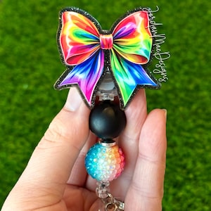 May include: A rainbow glitter bow with black glitter accents is attached to a black bead and a rainbow iridescent bead. The beads are attached to a retractable badge holder with a clear plastic clip.