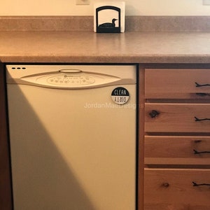 Peut inclure: Un lave-vaisselle blanc avec un aimant noir "Propre/Sale" fix&eacute; &agrave; la porte. Le lave-vaisselle est dans une cuisine avec des armoires en bois.