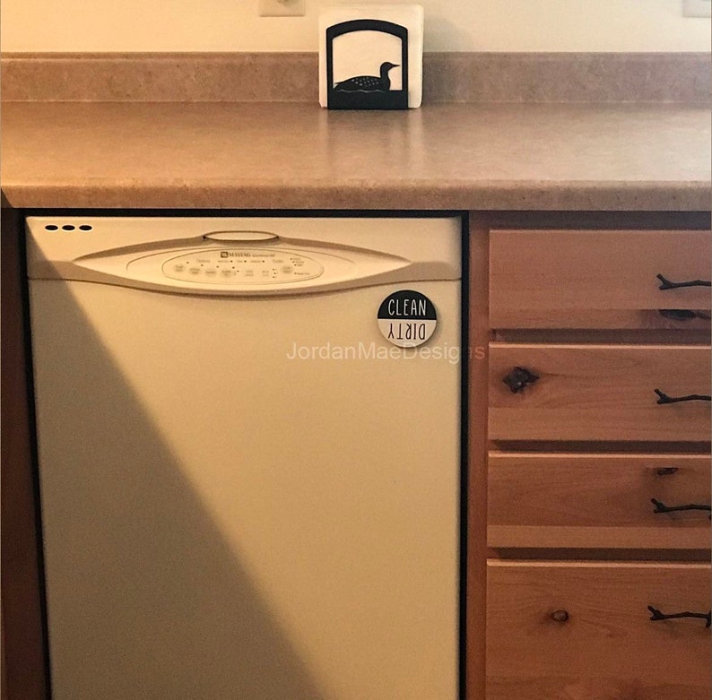 Peut inclure: Un lave-vaisselle blanc avec un aimant noir "Propre/Sale" fix&eacute; &agrave; la porte. Le lave-vaisselle est dans une cuisine avec des armoires en bois.