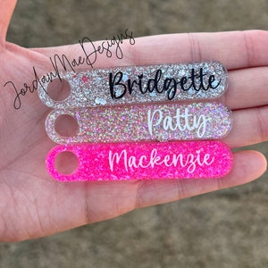 Puede incluir: Tres llaveros de purpurina rosa y blanca con los nombres "Bridgette", "Patty" y "Mackenzie" escritos en negro.