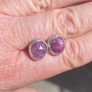 May include: A pair of round stud earrings with faceted, deep pink gemstones set in silver bezels. The earrings are displayed against a light-toned background.