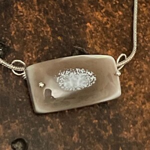 May include: A rectangular pendant necklace with a silver chain. The pendant is a light brown color with a white, oval-shaped design in the center. The necklace is a simple, elegant piece of jewelry.