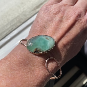 May include: A silver bracelet with a large, oval-shaped green gemstone. The bracelet is made of wire and has a simple, elegant design.