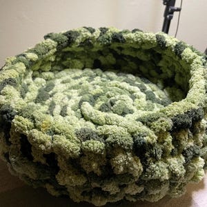 May include: A green and brown crocheted pet bed with a round shape. The bed is made of soft, fluffy yarn and has a cozy, inviting look.