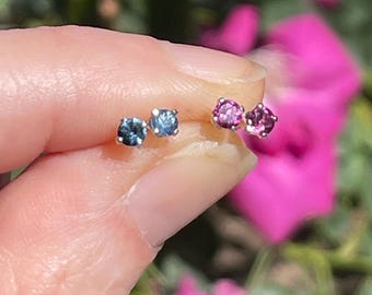May include: Two pairs of stud earrings. One pair features round, blue-green gemstones, and the other pair has round, pink gemstones. The earrings are set in silver or white gold. The background is blurred with pink flowers.