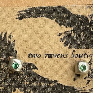 May include: A pair of silver earrings with small green gemstones set in a square shape. The earrings are on a brown background with the text "two ravens boutique".