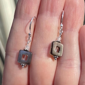 May include: Silver earrings with a square, open-centre design. Each earring has a small, clear bead and a cluster of tiny silver beads at the bottom of the square. The earrings are held by a person's hand.