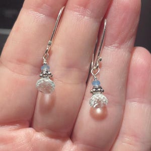 May include: A pair of dangle earrings with a silver hook and a clear, faceted, teardrop-shaped crystal. The earrings also feature a small, light blue bead and decorative silver accents. The earrings are shown against a blurred background.