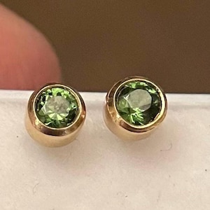 Puede incluir: Un par de pendientes de oro con piedras preciosas verdes. Los pendientes son redondos y tienen un diseño sencillo.