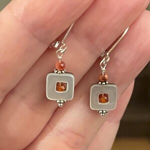 May include: Silver earrings featuring a square, pale grey frame with an orange gemstone centre. A small, round orange bead sits above the frame, and a cluster of small silver beads is below.