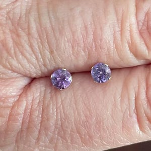 May include: A pair of round, light purple stud earrings. The earrings are set in a silver-colored metal and are displayed against a skin-toned background.