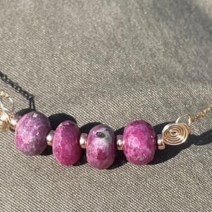 Op de afbeelding: Een delicate ketting met een gouden ketting en vijf gefacetteerde, dieproze robijnkralen. De ketting heeft aan elk uiteinde een spiraalvormig ontwerp. De ketting wordt gepresenteerd op een grijze ondergrond.
