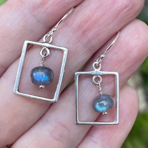 May include: A pair of silver earrings featuring a square frame design. Each earring has a small, round, iridescent blue gemstone suspended within the frame. The earrings are displayed against a natural background.