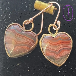 Puede incluir: Un par de pendientes en forma de corazón con un acabado en cobre. Los pendientes presentan una piedra de ágata roja, marrón y blanca con un patrón de remolino.
