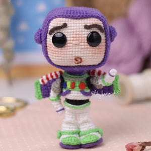 Op de afbeelding: Een gehaakt amigurumi-figuurtje van Buzz Lightyear met een grote paarse capuchon, zwarte plastic ogen en een gedetailleerd ruimtepak in wit, groen en paars. De tekst BUZZ LIGHTYEAR patrón de pago is zichtbaar in de linkerbovenhoek van de afbeelding.