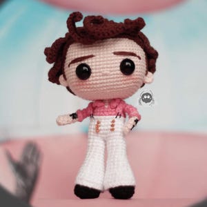 以下が含まれることがあります： 手作りのかぎ針編み人形。薄いピンク色のシャツ、白いズボン、濃い茶色の髪をしています。人形は黒い目を持ち、腕を横に添えてポーズをとっています。人形の衣装には金色のボタンが含まれています。人形の高さは約15cmです。