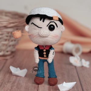 Puede incluir: Muñeco de ganchillo de un personaje de dibujos animados que lleva un gorro de marinero blanco, pelo negro y una camisa azul marino con cuello rojo. El muñeco tiene una pipa en la boca y está guiñando un ojo. El muñeco está junto a barcos de papel.