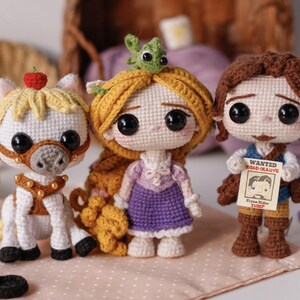 Op de afbeelding: Gehaakte poppen van een wit paard met een rode appel, een meisje met blond haar en een jongen met een "Wanted" poster. De poppen zijn handgemaakt met grote zwarte ogen en staan op een roze ondergrond.