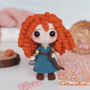 Puede incluir: Muñeca de ganchillo hecha a mano con cabello rizado naranja brillante, ojos negros grandes y un vestido azul. La muñeca sostiene un arco y una flecha marrones. La muñeca mide aproximadamente 15 cm de alto. Es un artículo hecho a mano.