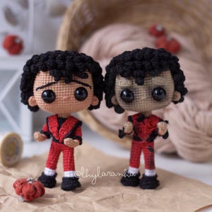 Puede incluir: Dos muñecos de ganchillo, cada uno representando una figura con un atuendo rojo con pelo y zapatos negros. Los muñecos tienen grandes ojos negros y están de pie. Una pequeña calabaza roja de ganchillo está en primer plano.