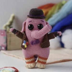 Puede incluir: Muñeca de elefante de ganchillo hecha a mano en rosa, con chaqueta y sombrero marrones. La muñeca tiene ojos negros grandes, pantalones a rayas naranjas y beige y una pajarita morada. Un pin colorido está adherido a la chaqueta. La muñeca está de pie con los brazos extendidos.
