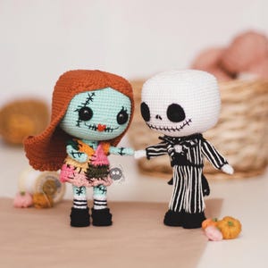 Puede incluir: Muñecos de ganchillo de Sally y Jack Skellington de Pesadilla antes de Navidad. Sally tiene la cara azul claro, pelo rojo y un vestido de retazos. Jack es blanco con cara de esqueleto y lleva un traje a rayas blanco y negro. Pequeñas calabazas a sus pies.