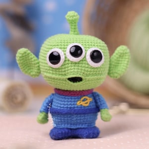 Puede incluir: Una pequeña muñeca de ganchillo hecha a mano de un alienígena de tres ojos con piel verde claro, grandes ojos negros y una antena. La figura lleva un traje espacial azul con un cuello morado y un emblema de planeta amarillo bordado en el pecho.