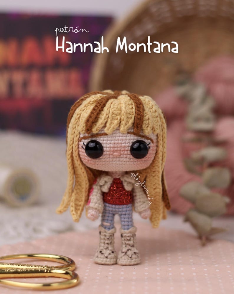 May include: Crocheted doll of Hannah Montana with blonde hair and a red sequined top, blue jeans, and beige boots. The doll has large black eyes and is handmade. The text "patr&oacute;n Hannah Montana" is visible in the background.