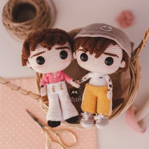May include: Two handmade crochet dolls, one in a pink shirt and white pants, the other in a white shirt and yellow shorts, holding hands. They have brown hair and large black eyes. The dolls are in a woven basket with a pair of scissors.