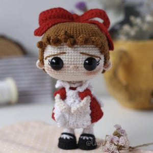 Könnte beinhalten: Handgefertigte Häkelpuppe mit roter Schleife, braunen Haaren und einem rot-weißen Kleid. Die Puppe hat große schwarze Augen und schwarze Schuhe. Die Puppe steht auf einer rosa Oberfläche.