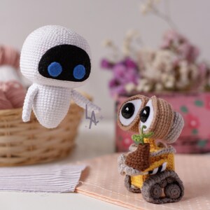 Puede incluir: Figuras de ganchillo de los personajes Eva y Wall-E de la película animada. Eva es blanca con cara negra y ojos azules. Wall-E es amarillo y marrón, sosteniendo una pequeña planta. Figuras hechas a mano.