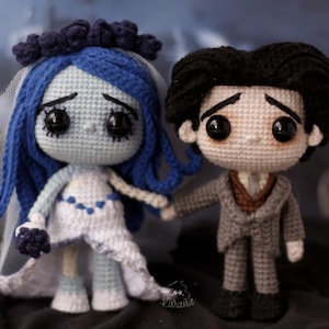 May include: Crocheted dolls of a bride and groom, inspired by a movie. The bride doll has blue hair, a white dress, and a veil. The groom doll wears a gray suit. Both dolls have large black eyes and are holding hands.