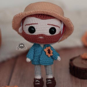 Könnte beinhalten: Gehäkelte Puppe, die eine Figur mit rotem Bart darstellt, die eine blaue Jacke, braune Schuhe und einen beigefarbenen Hut trägt. Die Puppe hat große schwarze Augen und eine Sonnenblumenbrosche. Die Puppe ist etwa 15 cm groß.
