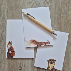 Può includere: Tre blocchi per appunti rettangolari in carta bianca con illustrazioni ad acquerello di una volpe, uno scoiattolo e un ghepardo. Una penna a sfera in legno chiaro con clip e punta argentate poggia in diagonale sul blocco superiore.