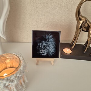 May include: A black and white image of a lion's face on a ceramic tile, displayed on a small wooden easel.