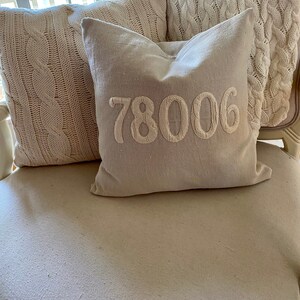 May include: A white armchair with three pillows. Two pillows are made of a white cable knit fabric. The third pillow is a light beige linen pillow with the number "78006" embroidered in white.