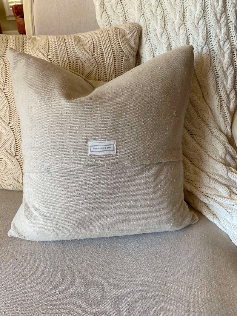 May include: A beige throw pillow with a white label that reads "The Stationery Shoppe". The pillow is made of a textured fabric and has a subtle sheen.
