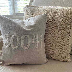 May include: Two decorative throw pillows on a white chair. One pillow is a cream-colored cable knit pillow. The other pillow is a light gray pillow with the number "30041" embroidered in white.