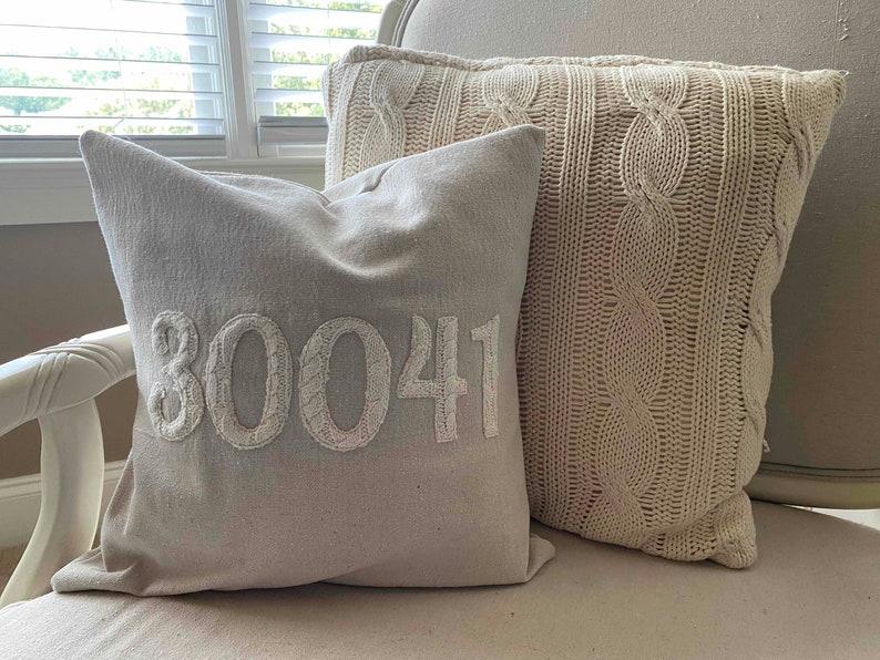 May include: Two decorative throw pillows on a white chair. One pillow is a cream-colored cable knit pillow. The other pillow is a light gray pillow with the number "30041" embroidered in white.