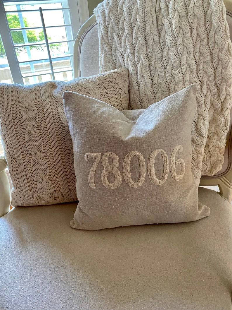 May include: A white armchair with three pillows. Two pillows are made of a white cable knit fabric. The third pillow is a light beige linen pillow with the number "78006" embroidered in white.