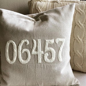 May include: A beige throw pillow with the number 06457 embroidered in white on the front. The pillow is sitting on a couch with a cable knit throw pillow beside it.