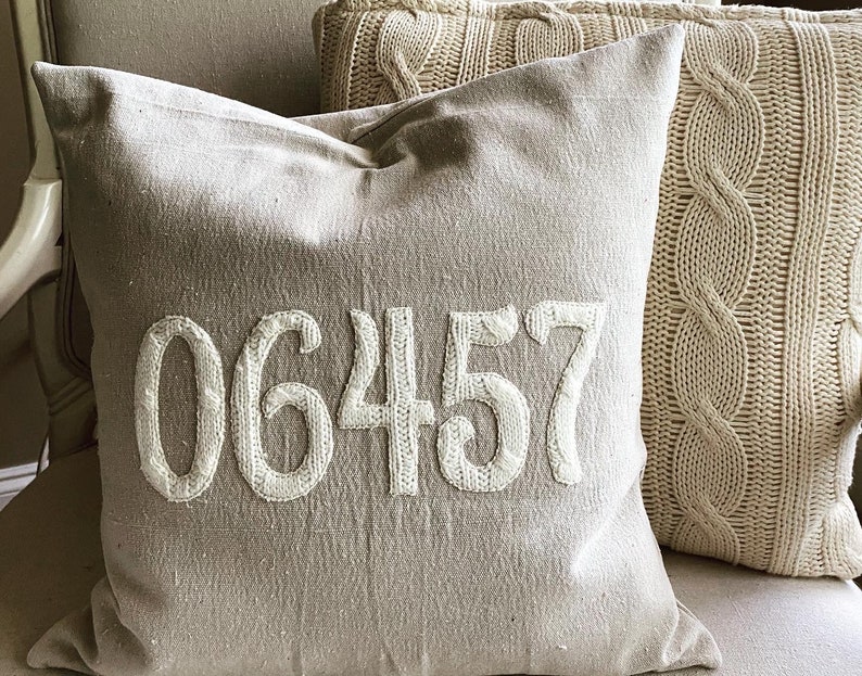 May include: A beige throw pillow with the number 06457 embroidered in white on the front. The pillow is sitting on a couch with a cable knit throw pillow beside it.