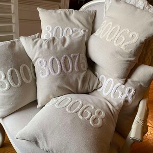 May include: A set of six beige throw pillows with white embroidered numbers, including 3000, 3002, 30075, 3062, 3068, and 3068. The pillows are arranged on a white armchair.