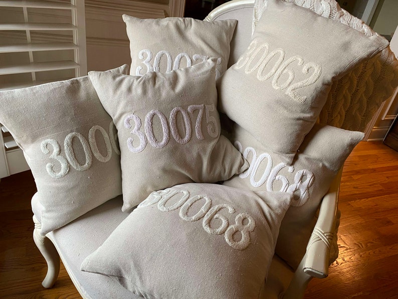 May include: A set of six beige throw pillows with white embroidered numbers, including 3000, 3002, 30075, 3062, 3068, and 3068. The pillows are arranged on a white armchair.