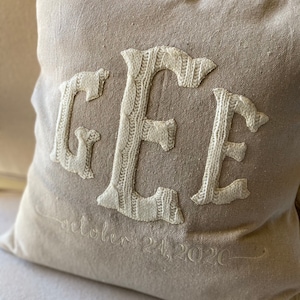 May include: A beige pillow with the letters "GEE" embroidered in white yarn. The letters are in a cable knit style. The date "October 21, 2020" is embroidered below the letters.
