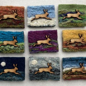 May include: Nine rectangular fiber art pieces arranged in a grid, each featuring a laser-cut wooden hare silhouette centered on a textured, multi-colored wool background. The backgrounds depict varied landscapes and skies in shades of blue, green, purple, orange, and yellow, with two pieces showing a small white circular moon.