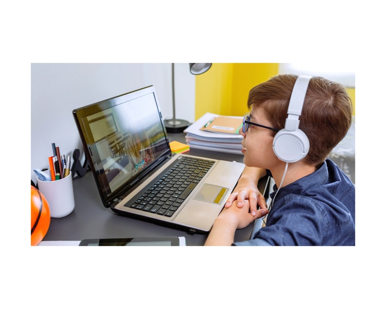 May include: A young person wearing headphones and glasses sits at a desk in front of a laptop computer. The laptop screen shows a video call with another person. The desk is cluttered with school supplies, including a basketball, pencils, and notebooks.