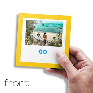 Puede incluir: Una tarjeta fotogr&aacute;fica con marco amarillo sostenida por una mano. La foto muestra a dos personas caminando hacia una playa con tablas de surf. La palabra "GO" est&aacute; impresa en azul en la tarjeta.
